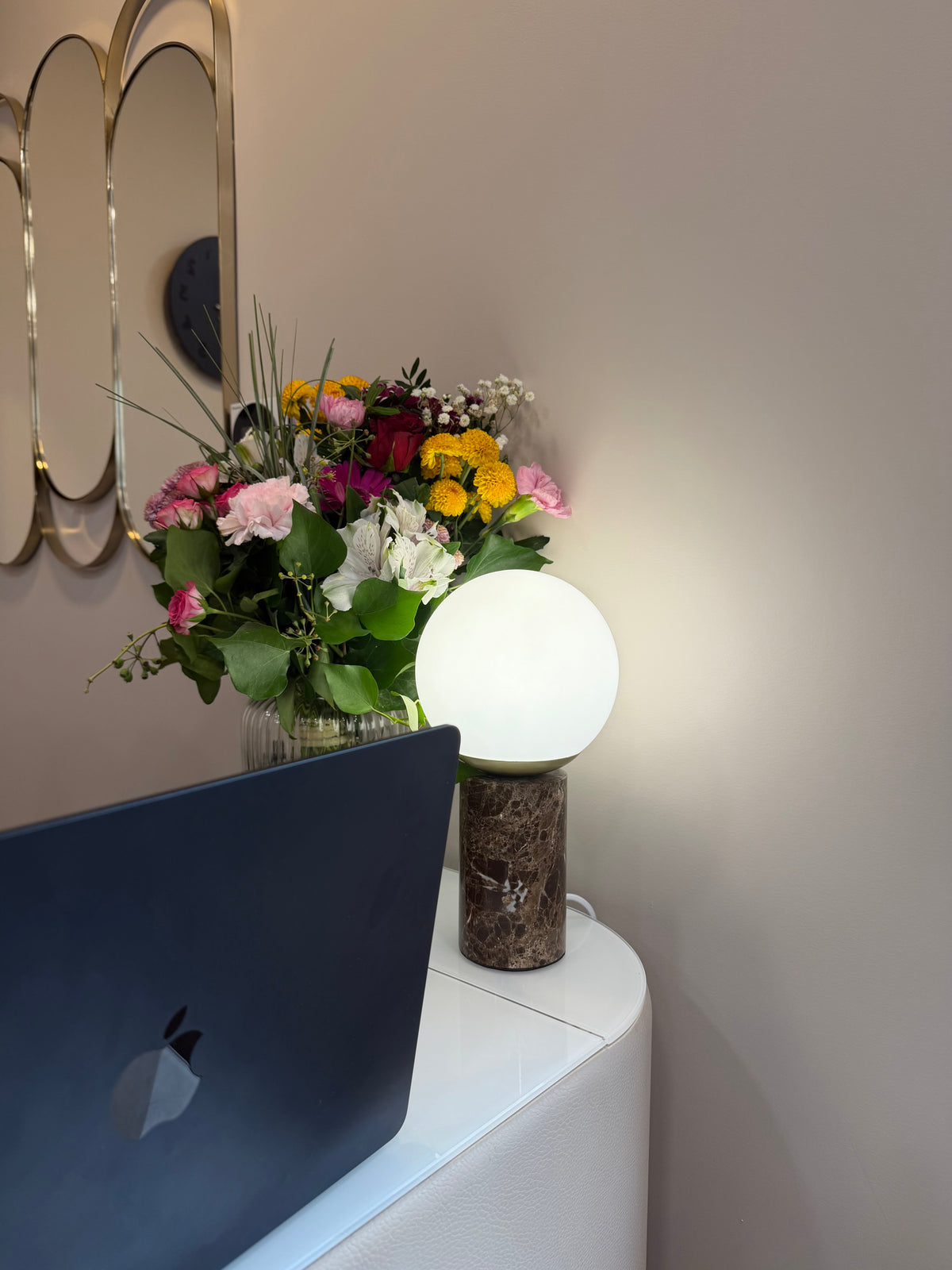 Bouquet de fleurs et lampe ronde posés sur un meuble blanc, devant un miroir doré